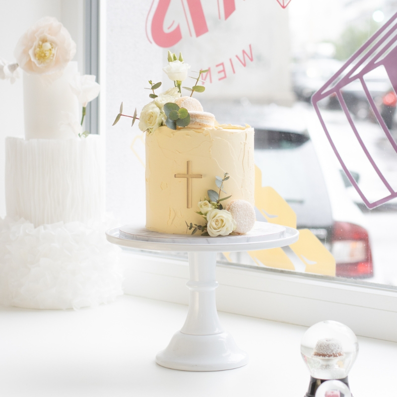 Eine zartgelbe Torte mit Blumen, Macarons und Kreuz auf einer Etagere.
